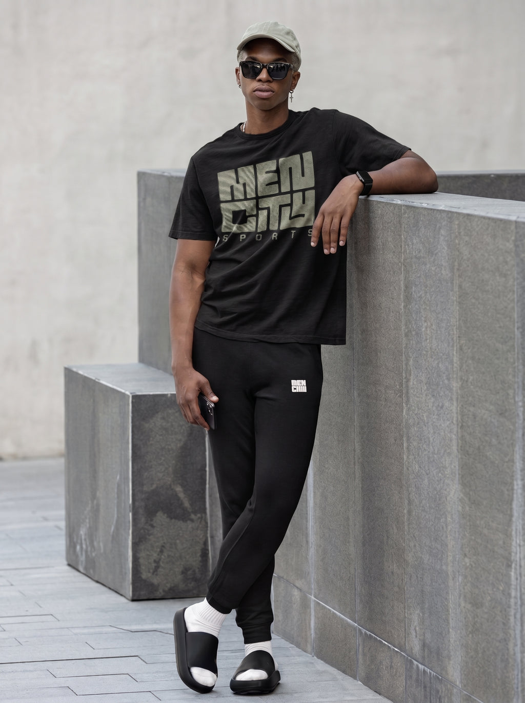 Man wearing black Mencity embroidered fleece gym joggers with a black tee, leaning on a concrete block