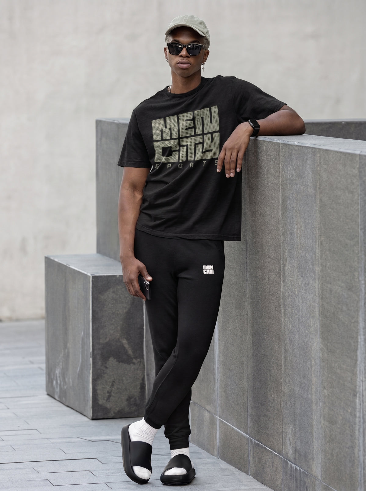 Man wearing black Mencity embroidered fleece gym joggers with a black tee, leaning on a concrete block